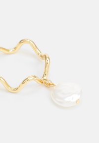 Guldvågig metallkedja med en blank vit pärla som hänge. Kedjan har en skinande yta och släta texturer som framhäver pärlans lyster.