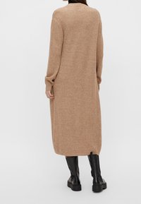 Langer brauner Strickcardigan mit geripptem Muster, langen Ärmeln und seitlichen Schlitzen, kombiniert mit schwarzen kniehohen Stiefeln.