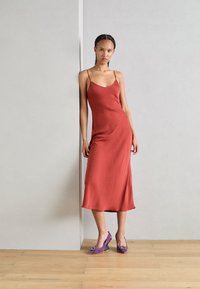 Abito a camicia in satin ruggine con spalline sottili, caratterizzato da una vestibilità rilassata e una silhouette dritta. Abbinato a tacchi viola decorati con abbellimenti.