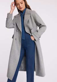 Kobieta w długim, szarym płaszczu zapinanym na podwójne guziki, nosząca niebieski dżinsowy kombinezon, z jedną ręką w kieszeni, stojąca na tle jasnego, jednolitego tła.