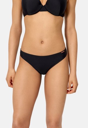 Mujer con un conjunto de bikini negro de diseño sencillo y liso, sobre un fondo claro y sencillo.