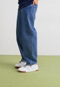 Kék farmer, bő szárú nadrág, elhalványult felülettel. Fehér sportcipővel párosítva, szürke részletekkel és texturált fűzőkkel.