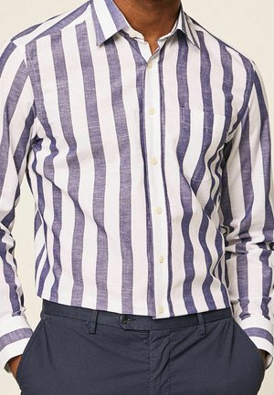 Homme portant une chemise blanche à manches longues avec des rayures verticales bleues et un pantalon bleu marine, mains dans les poches, recadré au niveau du cou et de la taille.