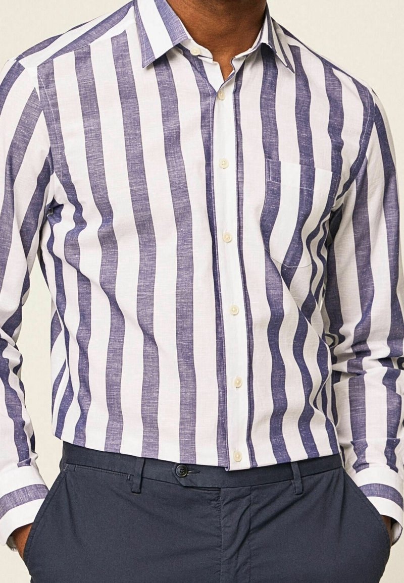 Homme portant une chemise blanche à manches longues avec des rayures verticales bleues et un pantalon bleu marine, mains dans les poches, recadré au niveau du cou et de la taille.