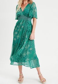 Robe maxi verte avec un col en V, des manches courtes volantées, un tissu à motifs et une taille élastique. Portée avec des sandales compensées couleur cuir tanné.