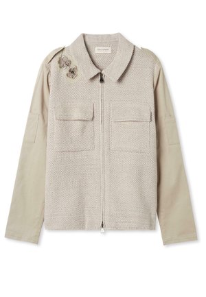 Veste beige clair avec devant en tricot, fermeture éclair, deux poches à rabat, manches en tissu lisse et papillons brodés décoratifs sur l'épaule.