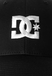 Gorra de béisbol negra con un gran logo bordado en blanco que presenta una estrella y letras estilizadas "DC" en la parte frontal. Diseño clásico y textura de tela.