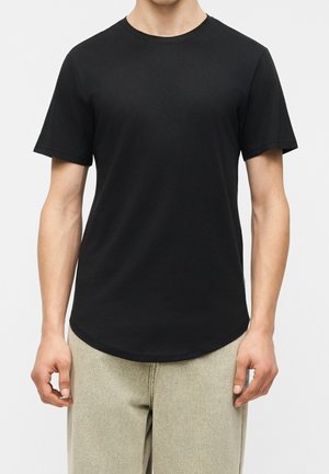 Homme portant un t-shirt noir uni à manches courtes et col rond et un pantalon beige clair, debout devant un fond blanc uni.