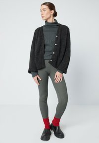 Črn puhast kardigan s srebrnimi gumbi, oblečen čez temno zeleno pulover s turtleneck izrezom. Olivno zeleni pajkice, rdeče nogavice in črne debele lesene cokle dopolnjujejo outfit.