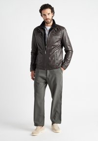 Bruine leren jas met een hoge kraag, een rits aan de voorkant en zijzakken. Gepaard met grijze broeken en beige instap schoenen.