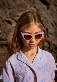 Ανοιχτό μωβ γυαλία ηλίου με κομμένες άκρες και βαθμιδωτούς καφέ φακούς. Φορεμένα με ένα λεβάντα πουκάμισο με μπλε κεντήματα και λεπτομέρειες από πεταλούδες.