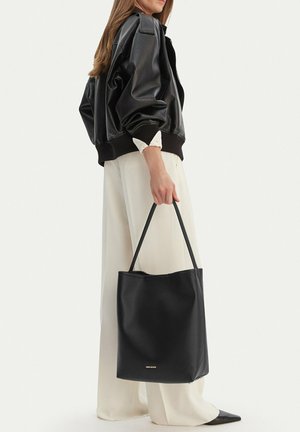 Kobieta w czarnej skórzanej kurtce i białych, szerokich spodniach, trzymająca duża czarną skórzaną torbę typu tote za uchwyty.