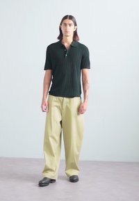 Polo negru cu textura, cu mâneci scurte, pantaloni bej lejere și pantofi negri tip slip-on. Design minimalist pe un fundal neutru.