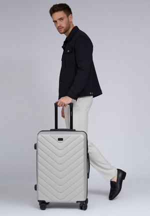 Hartschalenkoffer in Silber mit einem Chevron-Muster, schwarzem Trim, Doppelrädern und einem ausziehbaren Griff, der neben einer Person platziert ist.