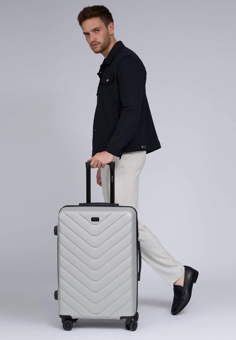 Hartschalenkoffer in Silber mit einem Chevron-Muster, schwarzem Trim, Doppelrädern und einem ausziehbaren Griff, der neben einer Person platziert ist.