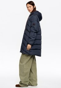Kobieta w granatowej, puchowej kurtce z kapturem, szerokich oliwkowozielonych spodniach i brązowych butach, stojąca bokiem na białym tle.