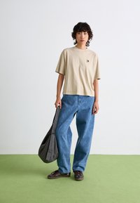T-shirt beige à manches courtes avec logo, assortie à un jean léger à jambes larges bleu clair, un sac gris à carreaux et des chaussures marron foncé sur un sol vert.