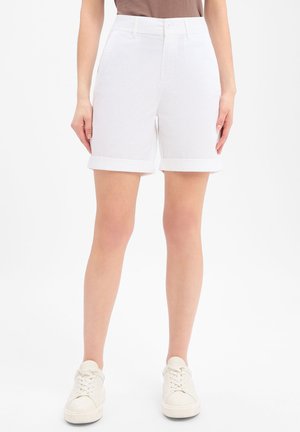 Vrouw draagt witte omslagshorts, beige top en witte sneakers, staand tegen een effen witte achtergrond.