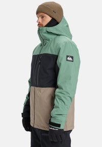 Multicolored jacket features a green and black top section, tan lower section, and hand pockets. Material is smooth, designed for outdoor use.