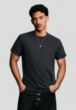 Young man with curly hair wearing a black rolled-sleeve t-shirt, dark pants with button details, and a pendant necklace, hands in pockets.