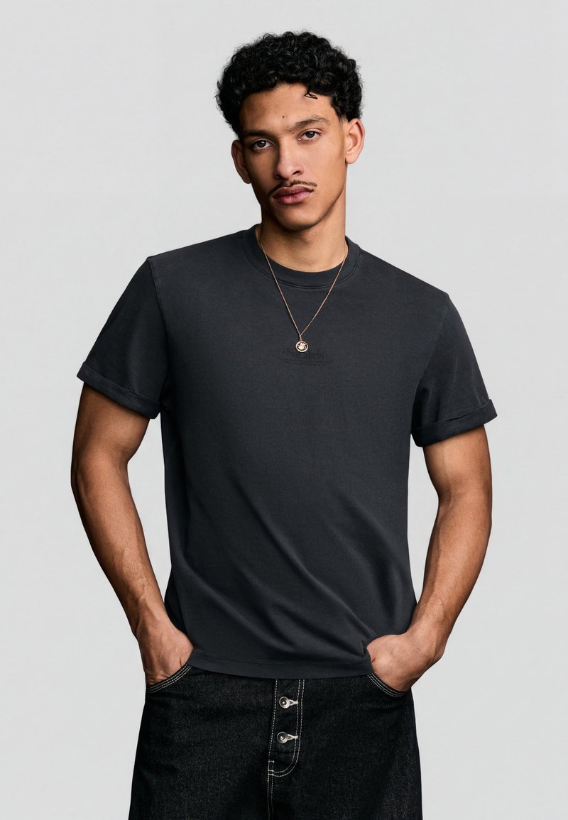 Jeune homme aux cheveux bouclés portant un t-shirt noir aux manches retroussées, un pantalon foncé avec des détails à boutons, et un collier pendentif, les mains dans les poches.