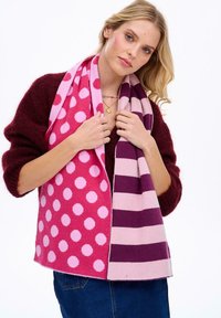 Ružová šatka s veľkými bielymi bodkami na jednej strane a striedavými pruhmi v svetlej a tmavej fialovej na druhej strane, vyrobená z mäkkej látky.