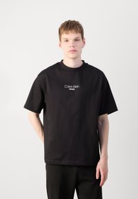 Crna pamučna majica kratkih rukava s okruglim izrezom. Na sredini se nalazi bijeli logo "Calvin Klein JEANS".