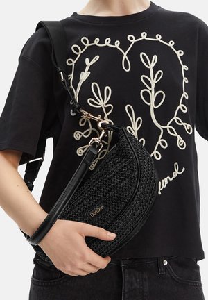 Persona con camiseta negra con bordado de enredadera blanca sostiene un bolso cruzado DeeZee negro tejido con tira de cuero sobre el pecho.