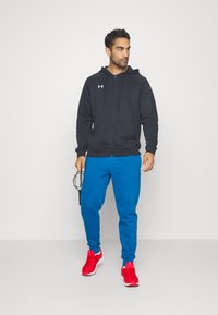 Svart zip-up hettegenser, blå joggebukser og røde trening-sko. Holder et hoppetau. Uformell atletisk bekledning med en avslappet passform.
