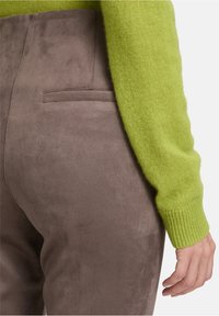 Close-up van een persoon die zachte bruine suède broek draagt met een achterzak en een felgroene gebreide mouw van een trui.