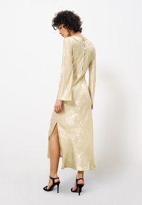 Langärmliges, metallisch goldenes Kleid mit einem Schlüsselloch-Ausschnitt im Rücken und Knopfverschluss. Mit einem Seitenschlitz und glatter Textur. Schwarze High-Heels.