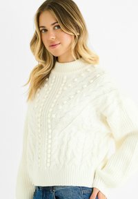 White cable-knit sweater featuring a high collar, textured patterns, and pom-pom accents, with ribbed cuffs and hem. Paired with denim.