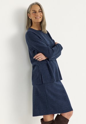 Vrouw met grijs haar in een blauwe gebreide trui en rok, glimlachend met gekruiste armen, leunend tegen een witte muur.