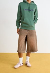 Zelená mikina s kapucí a fialovým logem, béžové denimu culottes a světle růžové tenisky s tmavými akcenty, na žlutém pozadí.