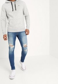 Šedá mikina s černými stahovacími šňůrkami, přední kapsa; kombinováno s roztrhanými modrými džíny s nášivkami a bílými teniskami.