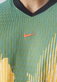 Kortärmad atletisk tröja i strukturerad blågrön tyg med svart v-ringning och orange Nike-logga på vänster bröst.