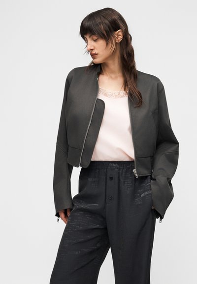 Γυναίκα με σκούρα μαλλιά φοράει ένα κοντό σκουρόχρωμο σακάκι πάνω από μια ανοιχτόχρωμη μπλούζα με δαντελωτή επένδυση και άνετο μαύρο παντελόνι με διακριτικά μοτίβα κειμένου.