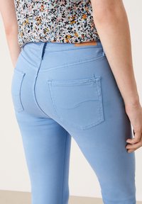 Jeans ajustados de color azul claro con dos bolsillos traseros, detalles de costura en contraste y una textura suave y elástica. Llevados con una blusa de estampado.