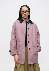 Παλτό με κόκκινο και λευκό καρό, καφέ γιακά από κορντόνι, μεγάλα κουμπιά και πλαϊνές τσέπες. Φορεμένο πάνω από ένα σκούρο πουλόβερ και πιετέ φούστα.