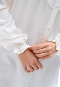 Biała bluzka z długimi rękawami, która ma elastyczne mankiety. Materiał wydaje się gładki i lekko przezroczysty, z guzikowym zapięciem z przodu. Ręce chwytają materiał.