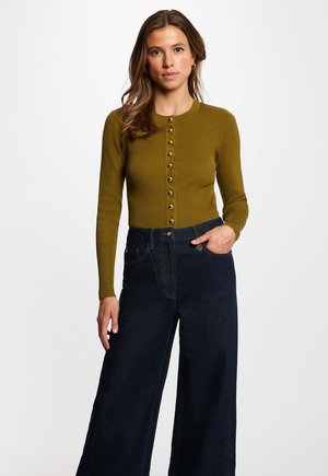 Vrouw met lang bruin haar die een olijfgroen geribd vest met knopen en donkere wijde jeans draagt, staand tegen een eenvoudige lichte achtergrond.