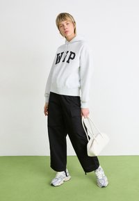 Grijze hoodie met "WIP" in zwarte letters, zwarte loszittende broek, witte tas en grijze sneakers. Soepele texturen en casual ontwerp.