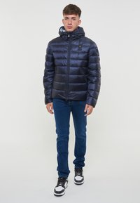 Giacca a vento blu navy con design trapuntato orizzontale, zip intera, cappuccio regolabile e polsini elasticizzati; abbinata a jeans blu e sneakers nere.