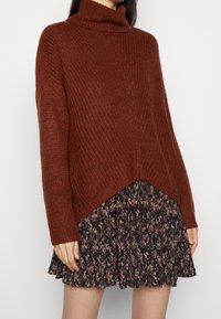 Knitted rust turtleneck sweater with a loose fit and ribbed texture, paired with a dark floral print flared dress, featuring a tiered design.