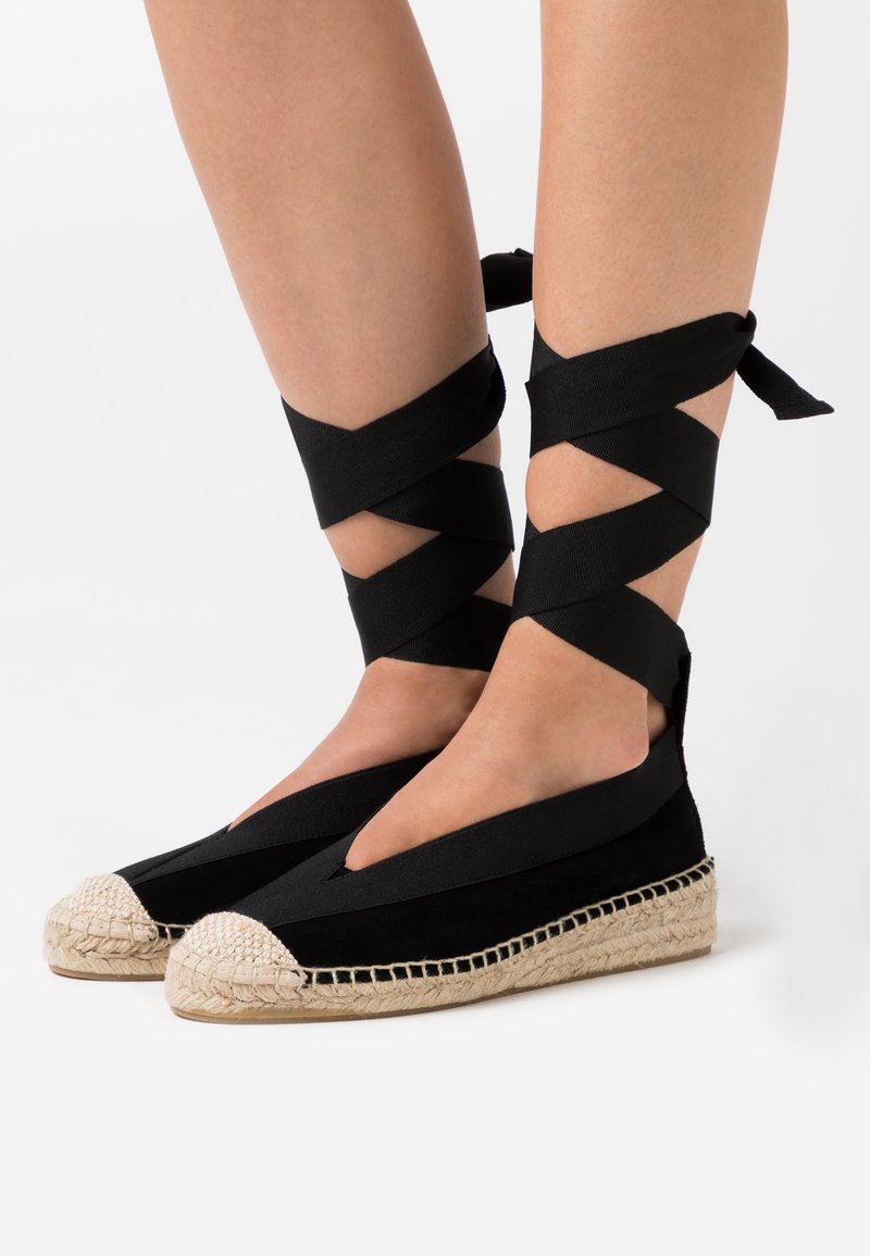Gambe che indossano espadrillas nere con punte beige intrecciate e cinturini alla caviglia avvolti intorno alle gambe inferiori su uno sfondo bianco.