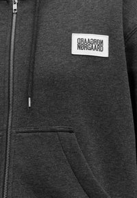 Sweat à capuche zippé gris foncé avec poche avant et écusson rectangulaire blanc avec texte noir en miroir « Nørgaard ».