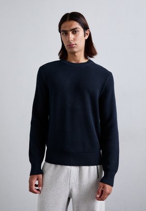 Suéter de punto azul oscuro con cuello y dobladillo acanalados, combinado con pantalones de chándal gris claro. Textura suave, diseño ajustado, mangas largas.