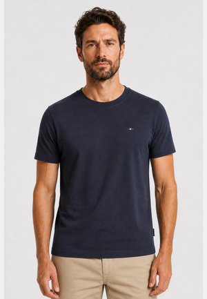 Mann mit kurzen dunklen Haaren und Bart, der ein einfarbiges dunkelblaues T-Shirt und beige Hose trägt, steht vor hellem Hintergrund.