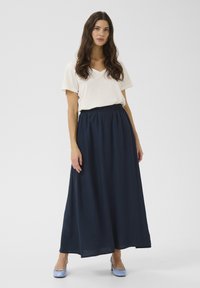 Jupe maxi plissée marine avec une taille élastique, associée à un t-shirt crème à col V ample et des chaussures à talons bleues.