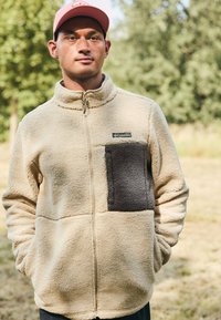 Beige fleece-takk med en grå lapp på venstre bryst, glidelås, høy krage og teksturert stoff. Uformelt yttertøy.